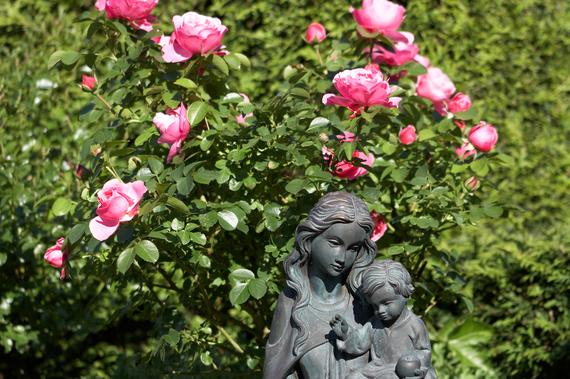 Bronzeskulptur einer Frau mit Kind vor einem Rosenstrauch mit pinken Blüten im Hintergrund.